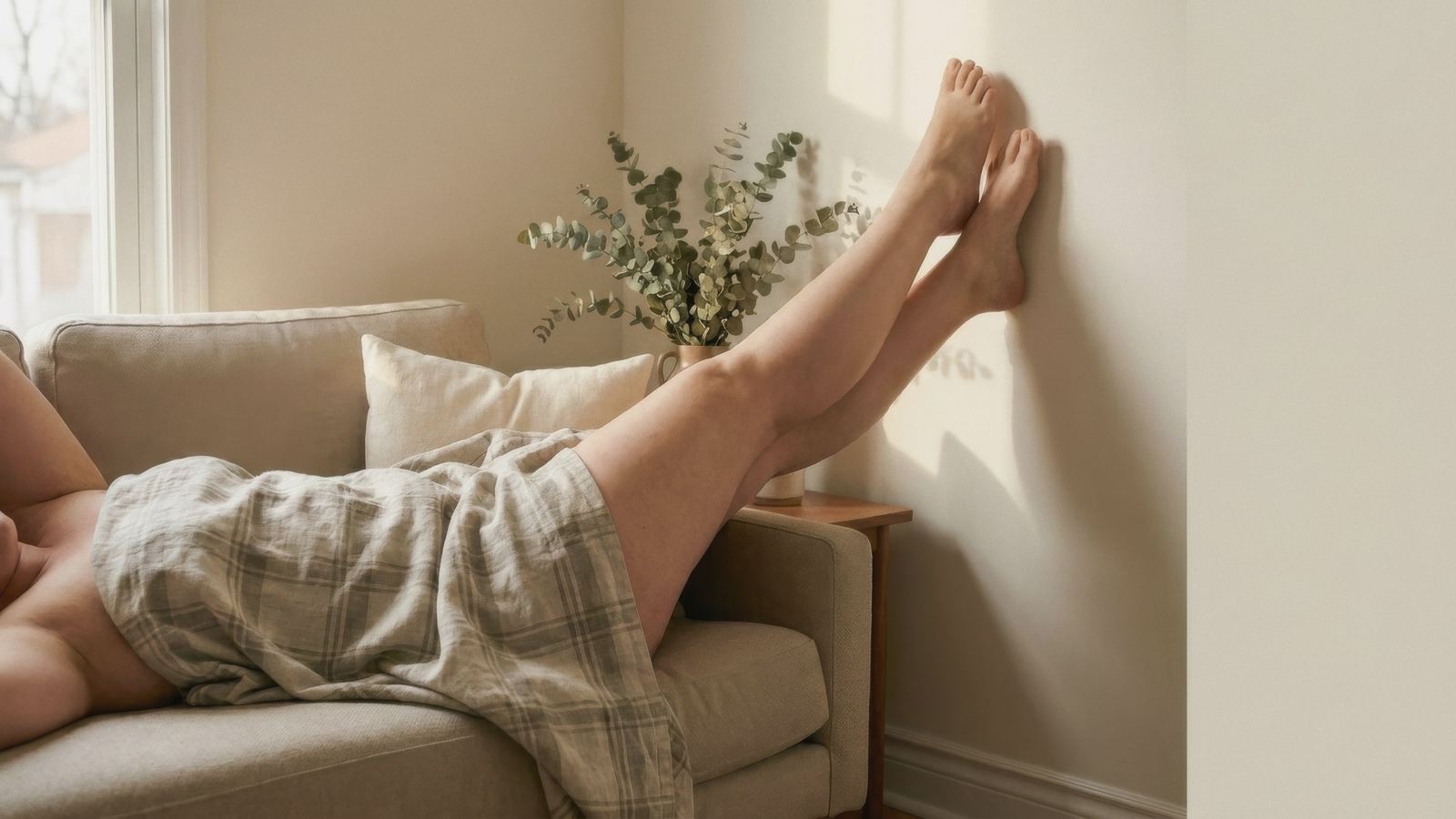 Une femme allongée les jambes surélevées contre un mur clair, dans un intérieur doux et lumineux — un moment de repos pour apaiser les jambes lourdes en fin de journée